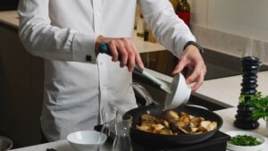 A Person Adds Sliced Garlic from A Small Bowl Into a Skillet with Golden-Brown Seared King Oyster Mushrooms