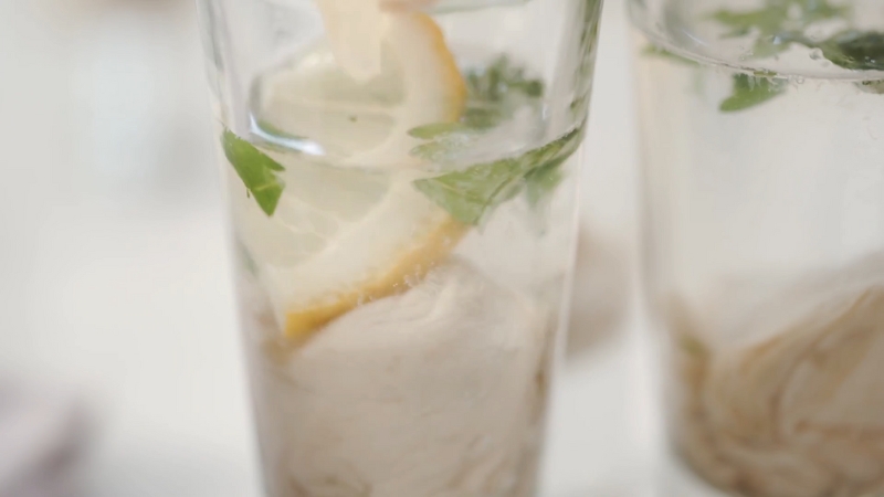 A Close-Up of An Oyster Shooter in A Glass with A Lemon Slice, Fresh Herbs, and A Clear Liquid