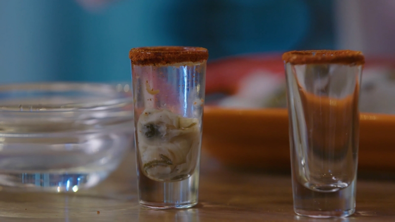 Two Shot Glasses Rimmed with Spice, One Filled with An Oyster and Clear Liquid, Sit on A Wooden Surface with A Blurred Background