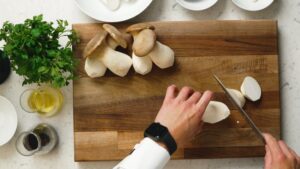 A Person Slices King Oyster Mushrooms on A Wooden Cutting Board with Fresh Parsley, Olive Oil, and Seasoning Nearby